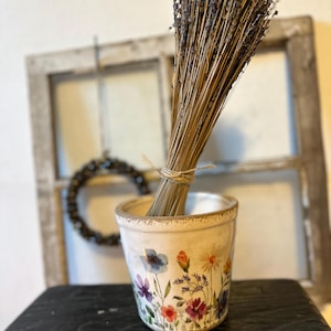Könnte beinhalten: Ein kleiner, cremefarbener Keramiktopf mit floralem Design, der ein Bündel getrockneten Lavendels enthält. Der Topf zeigt blaue, violette und rote Blumen. Der Lavendel ist mit Bindfaden gebunden. Der Topf steht auf einer dunklen, abgenutzten Holzoberfläche.