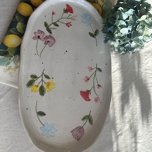 Op de afbeelding: Een ovale, crèmekleurige keramische schaal met handgeschilderde bloemmotieven in rood, geel, roze en blauw. De bloemen zijn gerangschikt in een delicaat, grillig patroon over het oppervlak. De schaal staat op een wit linnen tafelkleed.