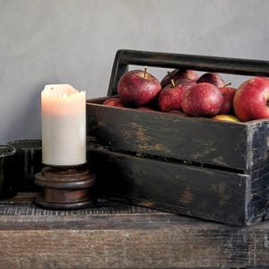 May include: A lit white pillar candle sits on a dark wooden base next to a weathered black wooden crate filled with several red apples. The crate features a handle and a distressed finish, while the candle emits a warm glow in the dim light.