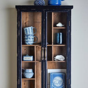 Könnte beinhalten: Ein schwarzer Holzschrank mit Glastüren, der verschiedene blaue und weiße Keramikartikel zeigt, darunter Tassen, einen Krug und dekorative Objekte. Der Schrank hat eine rustikale Oberfläche und Messinggriffe.