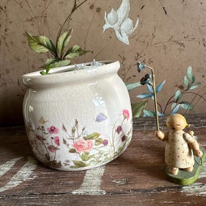 Può includere: Un vaso di ceramica color crema con un motivo floreale con rose rosa e farfalle viola. Una piccola statuina di legno di una ragazza che tiene una canna da pesca si trova accanto al vaso. Lo sfondo presenta una carta da parati floreale.