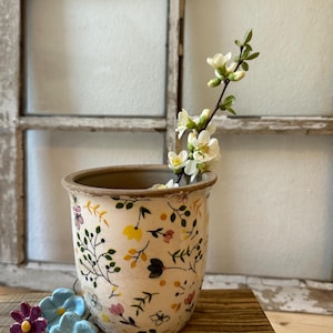 Puede incluir: Un macetero de cerámica blanca con un patrón floral, con pequeñas flores amarillas, rosas y verdes. El macetero está lleno de una rama de flores blancas.