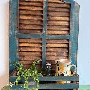 Könnte beinhalten: Rustikales, verwittertes Holzregal mit türkisblauem Rahmen und Lamellen aus Naturholz. Das Regal enthält eine Blumenvase, eine grüne Flasche und hängendes Grün. Das Design erinnert an einen Vintage- und Landhausstil.