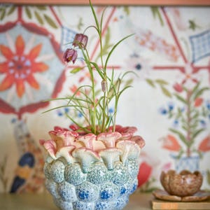 Op de afbeelding: Een unieke keramische bloempot met een textuur, paddenstoelachtig ontwerp in blauw, groen en roze tinten. De pot bevat een kleine plant met groene stengels en paarse bloemen. De achtergrond heeft een kleurrijk bloemenpatroon.