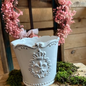 Op de afbeelding: Een witte keramische plantenbak met een verweerde afwerking, met een fleur-de-lis embleem en kroonontwerp. De plantenbak heeft een geschulpte rand en staat op een houten oppervlak met groen mos. Een roze bloemenkrans staat op de achtergrond.