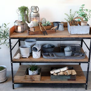 Op de afbeelding: Een metalen rek met drie niveaus met houten planken, waarop verschillende tuingereedschappen, plantenbakken en decoratieve items worden getoond. De planken zijn gemaakt van gerecycled hout en hebben een rustieke afwerking. Het zwarte metalen frame heeft een eenvoudig ontwerp.