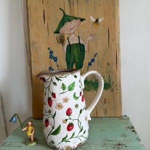 Op de afbeelding: Een witte keramische kan met een aardbei- en wijnstokpatroon. De kan heeft een bruine rand en een handvat. Hij staat op een groen houten oppervlak.