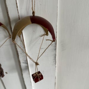 Puede incluir: Un adorno de metal rojo y dorado con forma de paracaídas con una pequeña caja de regalo roja colgando.