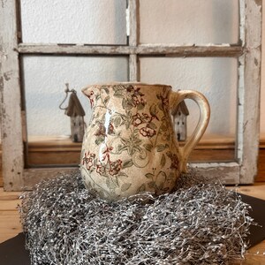 Könnte beinhalten: Eine beige Keramikkrug mit einem Blumenmuster in Rot und Grün. Der Krug hat einen gebogenen Griff und steht auf einem Bett aus silberfarbenen Zierstielen. Der Hintergrund umfasst einen verwitterten Fensterrahmen.
