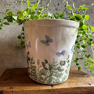 Może przedstawiać: Biała ceramiczna doniczka z błyszczącym, spękanym wykończeniem, ozdobiona nadrukiem zielonych liści botanicznych i małych niebieskich motyli wokół podstawy i boków, zawierająca zieloną roślinę z pnącymi się łodygami.