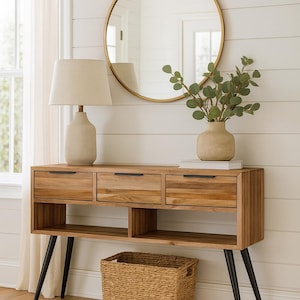 Op de afbeelding: Een houten console met drie lades en een onderste plank, met een beige lamp, een ronde gouden spiegel en een vaas met groen. Een geweven mand staat eronder. De tafel heeft zwarte poten.