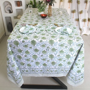 Op de afbeelding: Een wit tafelkleed met een groene en blauwe granaatappelprint. Het tafelkleed ligt op een tafel met twee couverts, elk met een wit bord, een groene servet en bestek.