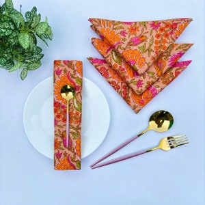 以下が含まれることがあります： ピンクとオレンジの花柄の布製ナプキン4枚セット。三角形に折り畳まれています。ナプキンは白い表面に置かれ、金色のスプーンとフォークが添えられています。