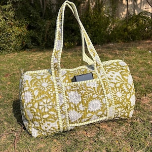 Peut inclure: Un sac de voyage en toile de jute à imprimé floral vert et blanc avec une poche zippée sur le devant. Le sac est bordé d'une bande blanche et verte.