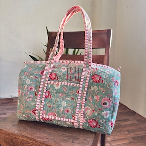 May include: A quilted duffel bag with a floral pattern in shades of teal, pink, and red. The bag has a rectangular shape with a zippered top and two handles. The handles and trim are striped in pink and white. The bag is sitting on a wooden chair.