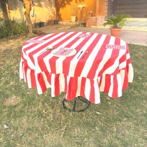 May include: A circular table featuring a red and white striped tablecloth with a frilled edge. A plate with a napkin and cutlery is set on the table. The table is positioned on a grassy lawn.