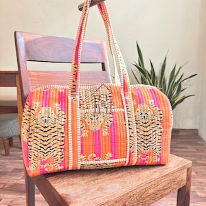 Puede incluir: Bolso tote acolchado con estampado de tigre. El bolso presenta rayas verticales naranjas, rosas y blancas, con ilustraciones de tigres en la parte delantera. El bolso tiene dos asas largas y un cierre de cremallera. El bolso está sobre una silla de madera.