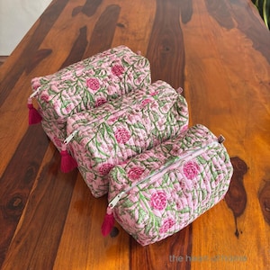 Könnte beinhalten: Drei rosa gesteppte Kosmetiktaschen mit einem Blumenmuster aus rosa Blüten und grünen Blättern. Jede Tasche hat einen Reißverschluss und eine rosa Quaste. Die Taschen sind auf einer Holzoberfläche angeordnet.