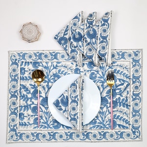 Può includere: Un'apparecchiatura con un piatto bianco, cucchiaio e forchetta dorati e tovaglietta e tovaglioli con motivo floreale blu. È presente anche un sottobicchiere in legno con un design circolare.