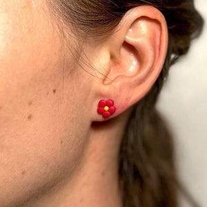 Op de afbeelding: Een paar rode bloem oorbel studs met gele centra. De oorbellen worden gedragen in de oorlel van een persoon.