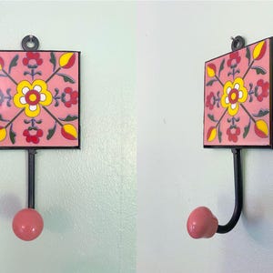 Könnte beinhalten: Dekorativer Wandhaken mit quadratischer, rosa Fliese mit floralem Design in Gelb, Rot und Schwarz. Der Haken hat einen schwarzen Metallschaft und einen rosa, abgerundeten Knauf. Er ist an einer hellgrünen Wand befestigt.