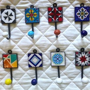 Könnte beinhalten: Acht dekorative Wandhaken mit bunten Keramikfliesen. Jeder Haken hat ein anderes Fliesendesign mit geometrischen Mustern und leuchtenden Farben wie Blau, Rot, Gelb und Grün.