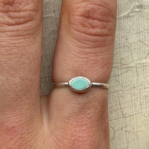 May include: A silver ring with a small, oval-shaped, turquoise stone set in the center. The ring is shown on a finger against a neutral background.