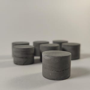 May include: A collection of six small, cylindrical objects made of dark gray concrete. Each piece features a smooth, flat top surface and a solid, uniform circular base, arranged in a cluster with one piece positioned in the foreground.