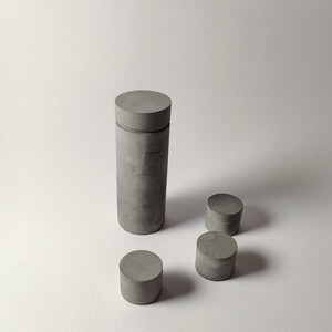 May include: A tall cylindrical grey concrete container with a matching lid, accompanied by three smaller, separate cylindrical grey concrete pieces of varying heights, all featuring a smooth, matte, and uniform grey finish throughout.