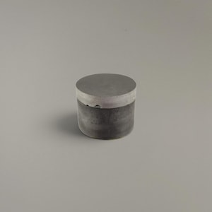 Könnte beinhalten: Ein kleiner zylindrischer Behälter mit zweifarbigem Design. Der obere Deckel ist hellgrau, während die Basis in einem dunkleren Anthrazit gehalten ist. Das Objekt hat ein glattes, massives Erscheinungsbild mit flacher Oberseite und geraden Seiten.