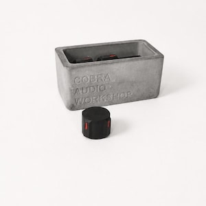 May include: A rectangular grey concrete enclosure with the embossed text COBRA AUDIO WORKSHOP on the front face. The interior contains two black switches with red accents. A separate black cylindrical knob with a red indicator line sits in front.