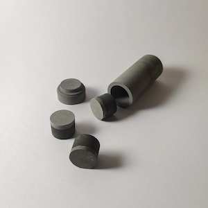 May include: A set of dark grey cylindrical objects including one hollow tube container, one stepped lid, and two solid circular discs. The items have a matte finish and a smooth texture, arranged in a cluster on a plain, light-coloured surface.