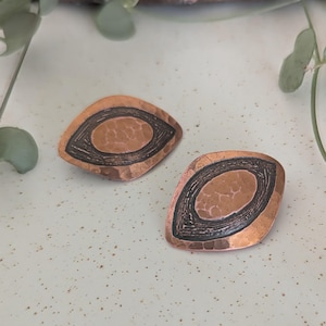 May include: A pair of copper-toned earrings with a hammered texture. Each earring has a dark, oval-shaped center framed by a black border. The earrings have a unique, geometric design.