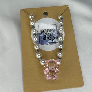 May include: A necklace featuring a pink teddy bear pendant. The necklace is made of white faux pearls and gray beads. The pendant is a translucent pink color. The necklace is displayed on a brown card with the text "pansy pop shop".