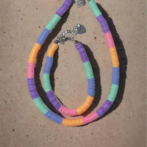 Può includere: Due collane di perline colorate con un ciondolo a forma di cuore. Le collane sono fatte di perline color arancio pastello, rosa, viola e verde. Le collane sono su uno sfondo sabbioso.