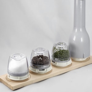 May include: A set of four clear plastic spice grinders with a gray electric grinder on a wooden base. The grinders contain salt, peppercorns, dried herbs, and a fourth grinder is empty.