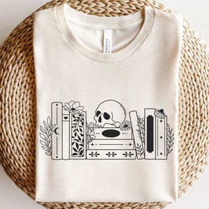 以下が含まれることがあります： 白いTシャツに、黒と白の骸骨と本のイラストが描かれています。本は積み重ねられており、骸骨は本の頂上に置かれています。花柄のアクセントが施されています。