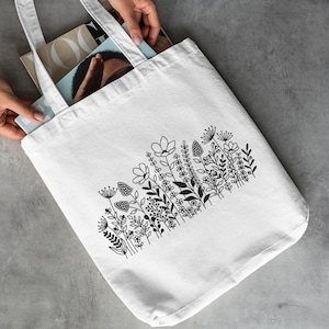 May include: White canvas tote bag featuring a black floral design. The design includes a variety of flowers and leaves in a simple, minimalist style.