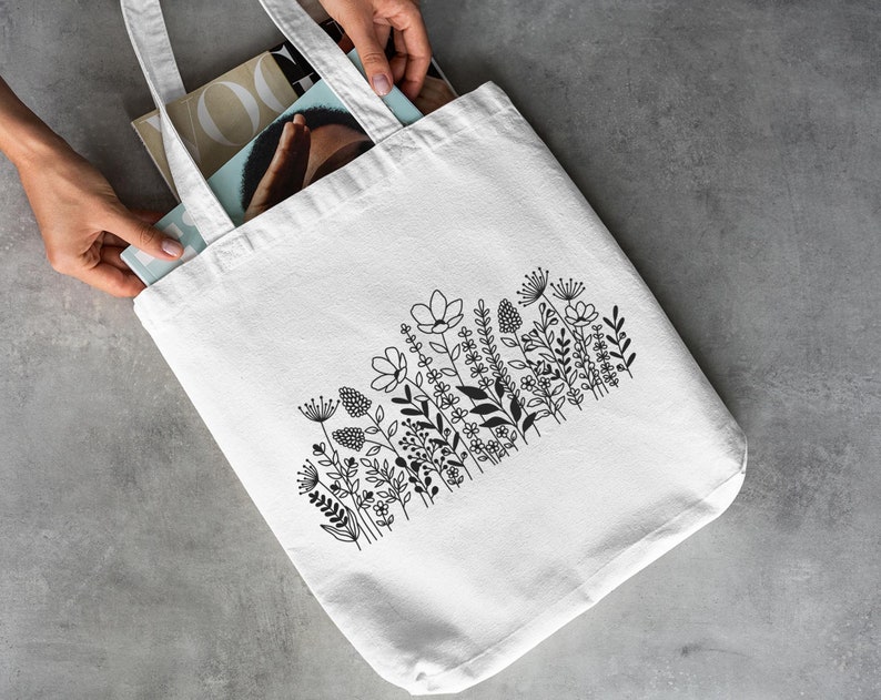 May include: White canvas tote bag featuring a black floral design. The design includes a variety of flowers and leaves in a simple, minimalist style.
