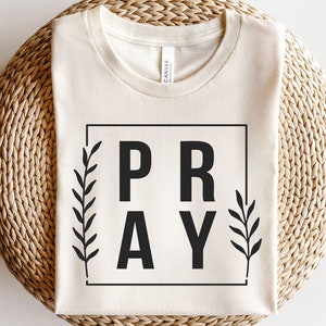 May include: A beige t-shirt with the word "PRAY" in a black square frame with black floral accents on each side.