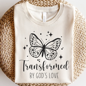 Può includere: Una maglietta beige con una grafica di farfalla nera e il testo "Trasformato dall'amore di Dio".