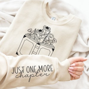 May include: A beige sweatshirt with a black graphic of an open book with flowers growing out of it. The text "JUST ONE MORE chapter" is printed on the sleeve.