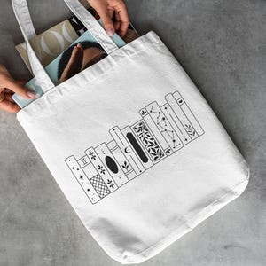 May include: A white tote bag with a black line drawing of a bookshelf. The bookshelf design features various decorative elements, including stars, moons, and floral patterns. The bag is held by hands and contains magazines.