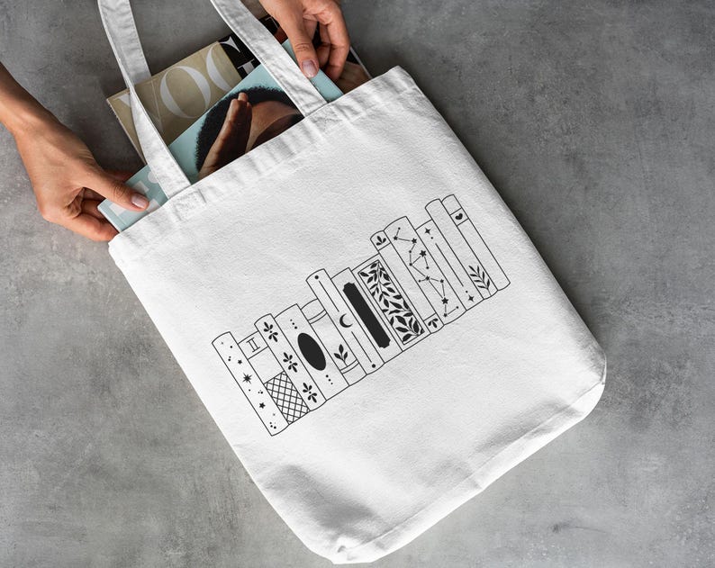 May include: A white tote bag with a black line drawing of a bookshelf. The bookshelf design features various decorative elements, including stars, moons, and floral patterns. The bag is held by hands and contains magazines.
