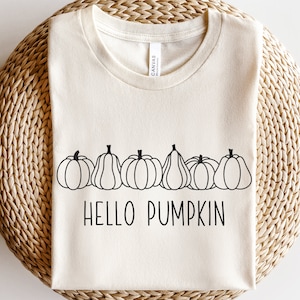May include: A beige t-shirt with a black outline of a row of pumpkins and the text "HELLO PUMPKIN".