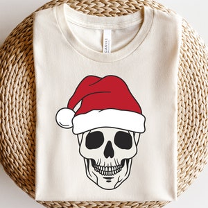 Puede incluir: Una camiseta blanca con una calavera en blanco y negro que lleva un gorro de Papá Noel rojo.