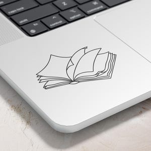 May include: A black and white line drawing of an open book with several pages turned. The book is on a white laptop, with the keyboard visible in the background. The image is a sticker or decal.