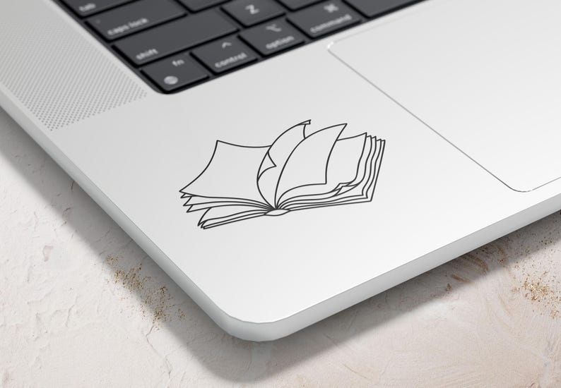 May include: A black and white line drawing of an open book with several pages turned. The book is on a white laptop, with the keyboard visible in the background. The image is a sticker or decal.