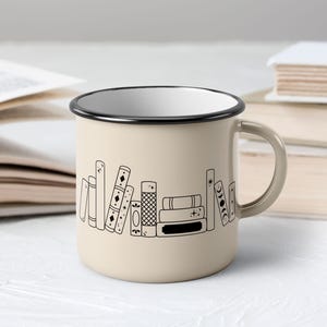 May include: Beige enamel mug with a black rim and handle. The mug features a black line drawing of a stack of books. The mug is sitting on a white surface with open books in the background.