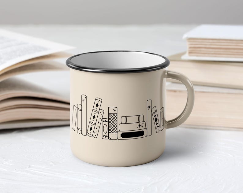 May include: Beige enamel mug with a black rim and handle. The mug features a black line drawing of a stack of books. The mug is sitting on a white surface with open books in the background.